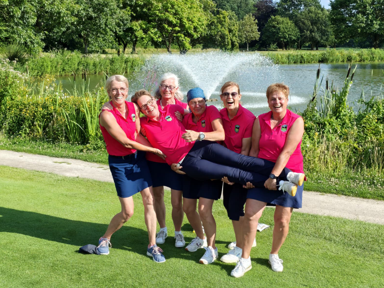 Golfclub Schmallenberg Damen AK50 Leverkusen Titel Die Damen der AK50 des Golfclub Schmallenberg in pinken Trikots und dunkelblauen Röcken oder Hosen halten lachend eine Spielerin quer vor sich hoch, im Hintergrund ein Teich mit einem Brunnen auf dem Golfplatz Leverkusen