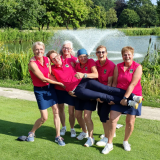 Die Damen der AK50 des Golfclub Schmallenberg in pinken Trikots und dunkelblauen Röcken oder Hosen halten lachend eine Spielerin quer vor sich hoch, im Hintergrund ein Teich mit einem Brunnen auf dem Golfplatz Leverkusen